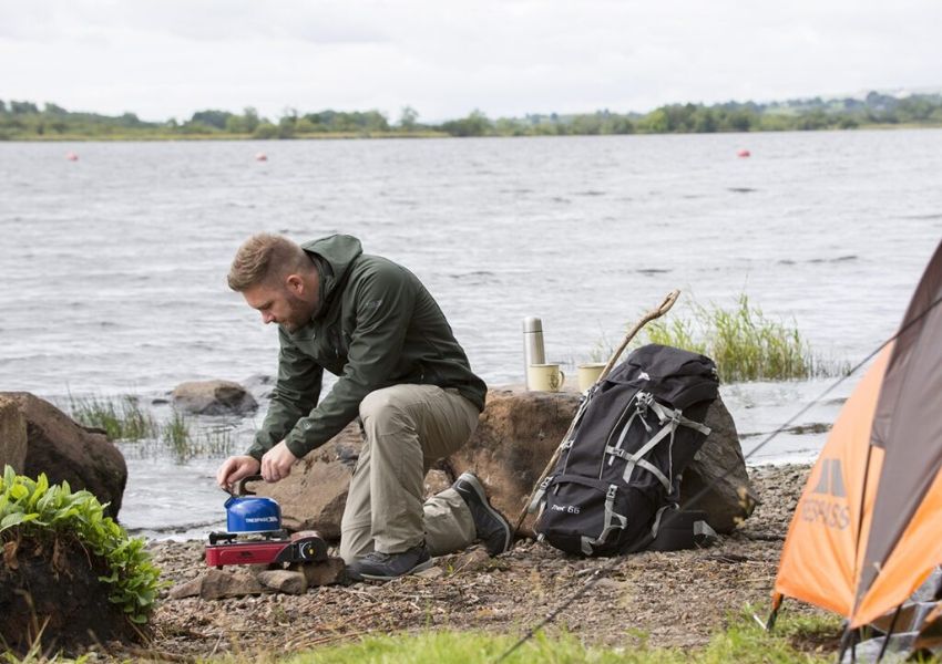 A man kneels by a lake, preparing a kettle on a camping stove next to a tent and backpack.