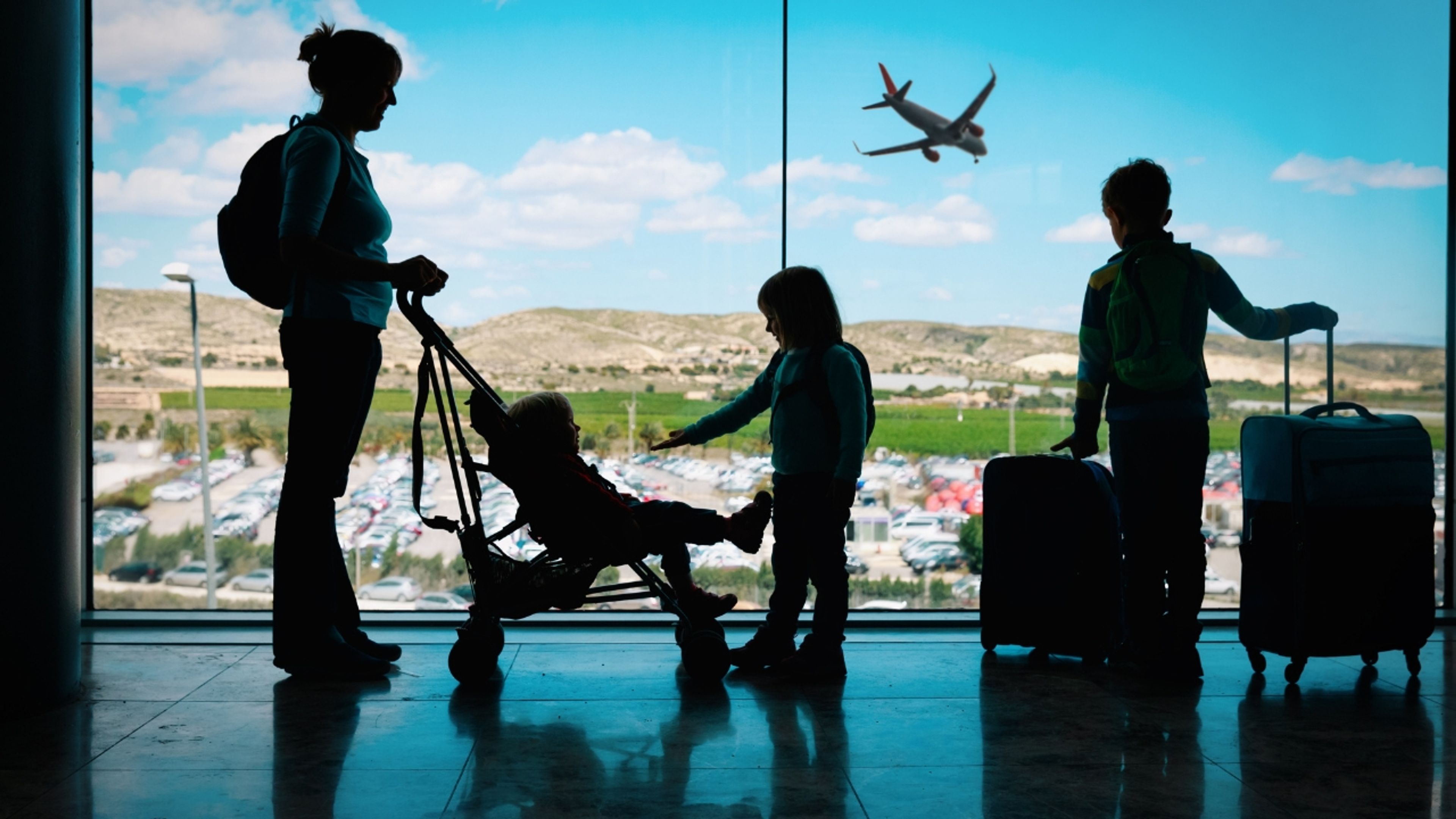 Silhouetted family with stroller and luggage by an airport window, an airplane flying outside.