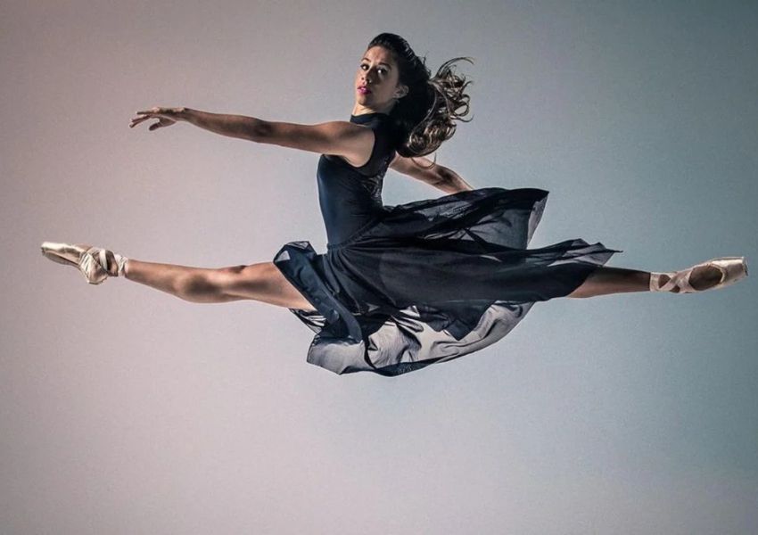 A ballet dancer in a black dress and pointe shoes leaps mid-air in a horizontal split.
