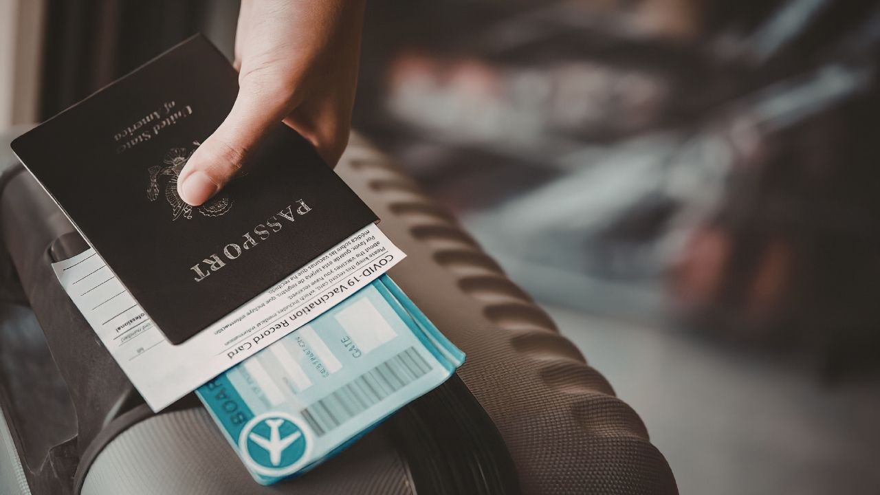 A hand holds a US passport, a COVID-19 vaccination record card, and a boarding pass over a suitcase.