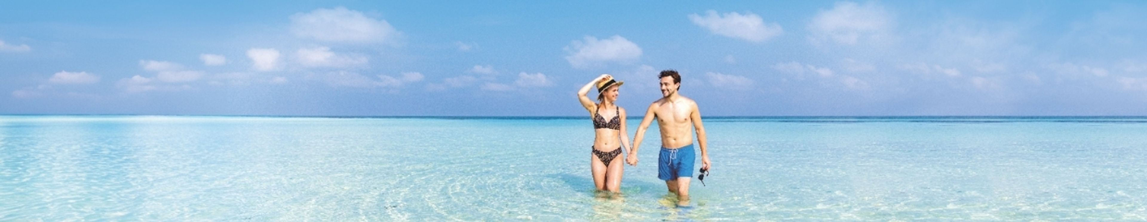 A couple walking hand-in-hand in shallow, clear blue ocean water.