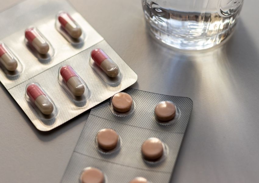 Blister packs of pink and beige capsules and round tablets next to a glass of water.