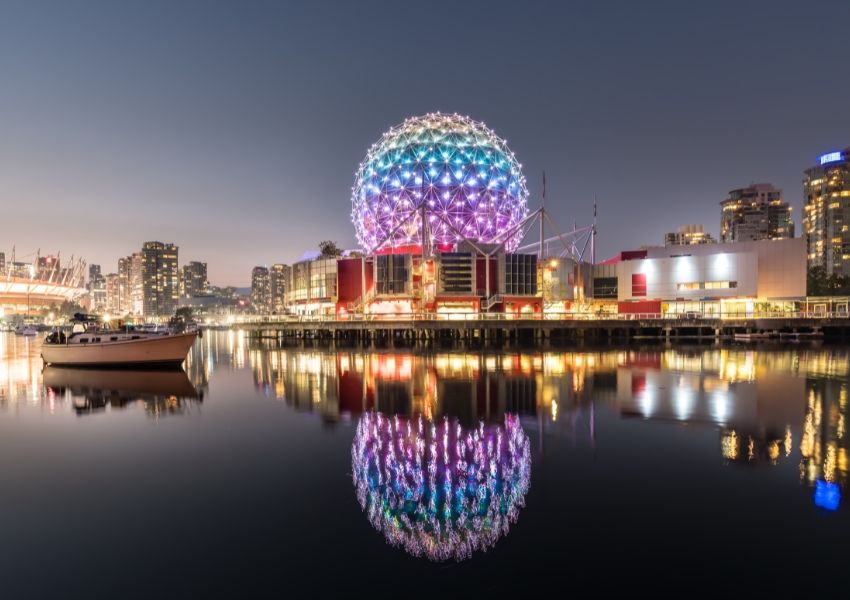 Colorful geodesic dome illuminated at night, reflected in calm water with a boat and city skyline.