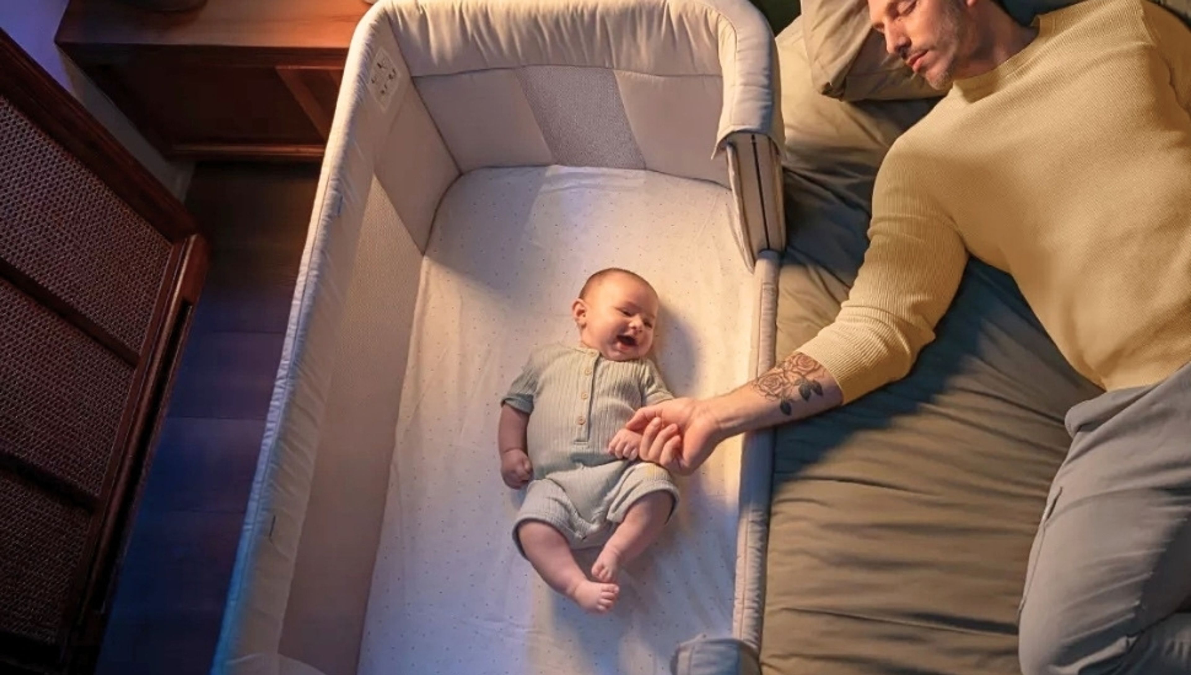 A sleeping man in bed holds the hand of a baby in an adjacent bassinet.