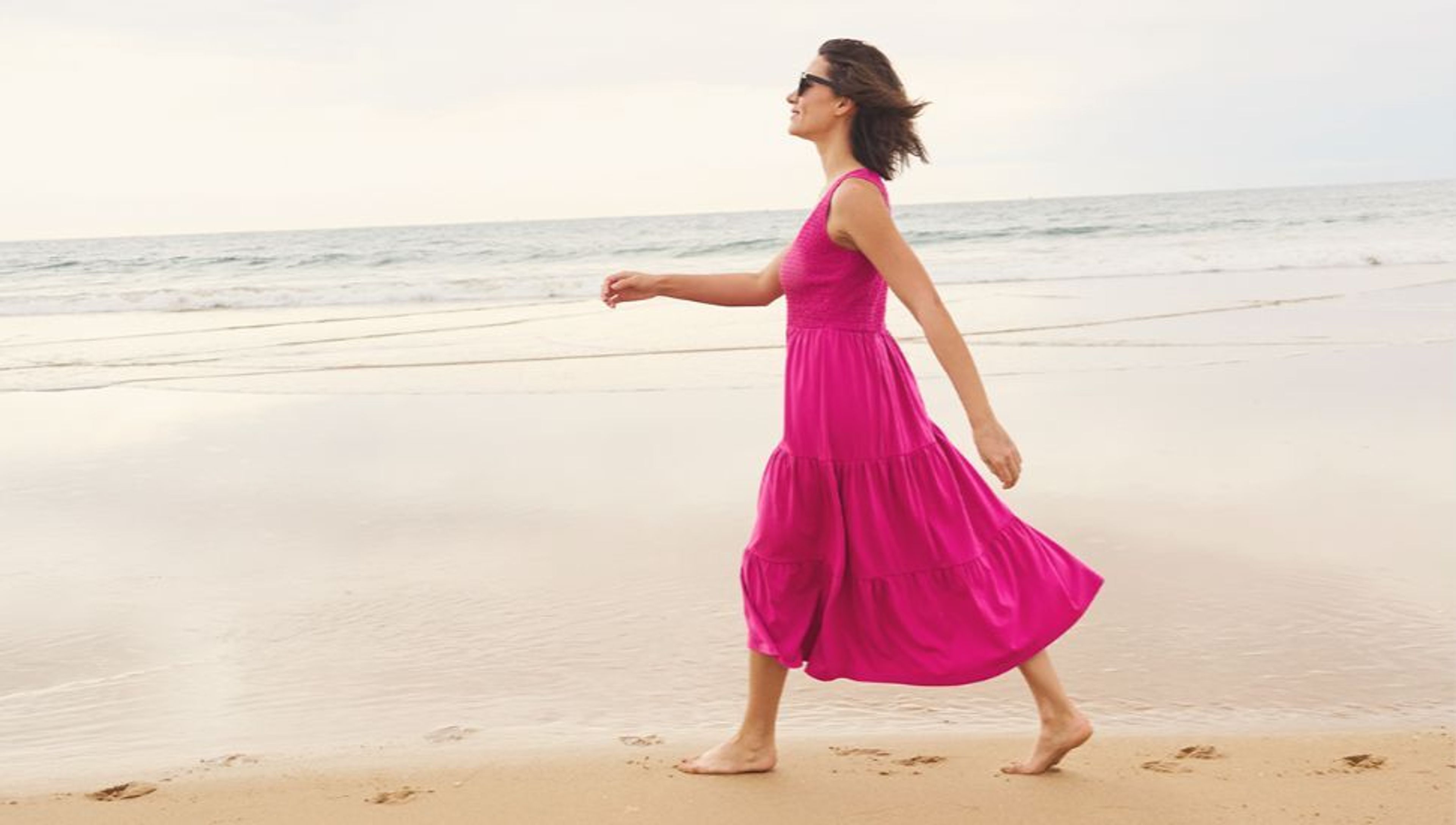 Woman in a bright pink dress walking barefoot on the beach.