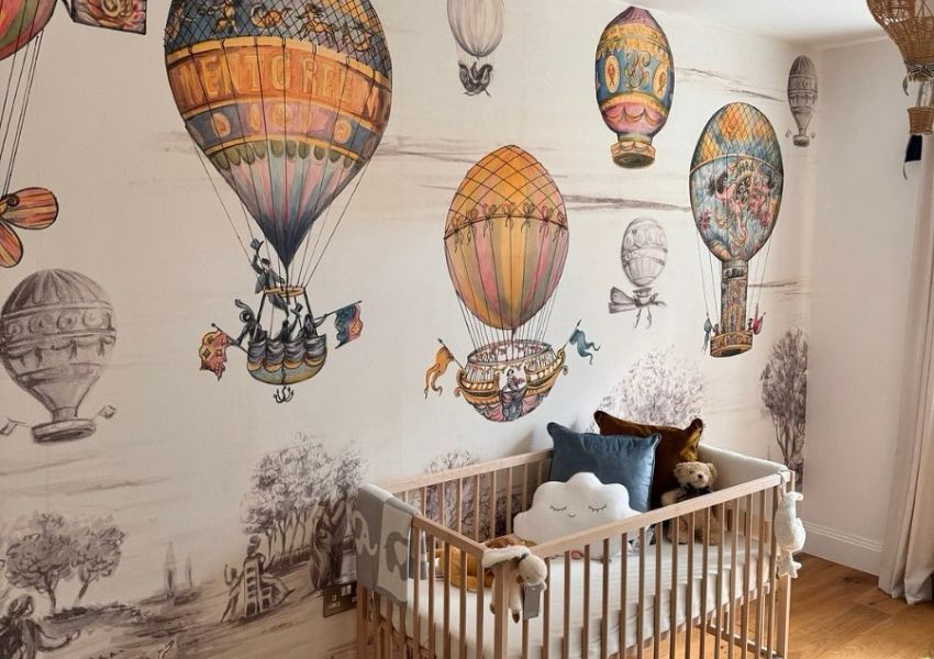 A child's room with a vibrant, vintage-style hot air balloon mural and a light wood crib filled with pillows and stuffed animals.