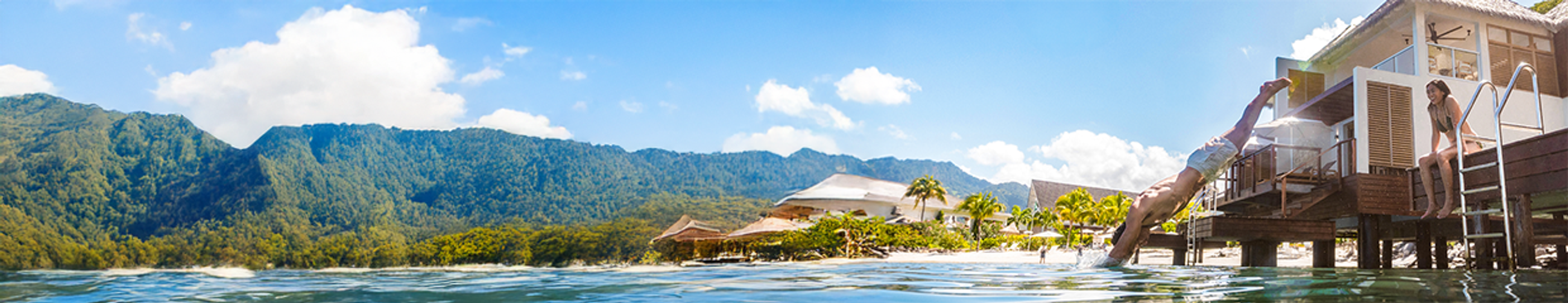 Panoramic view of a tropical overwater resort with lush mountains. A man dives into the ocean from a bungalow, while a woman sits on adjacent steps.
