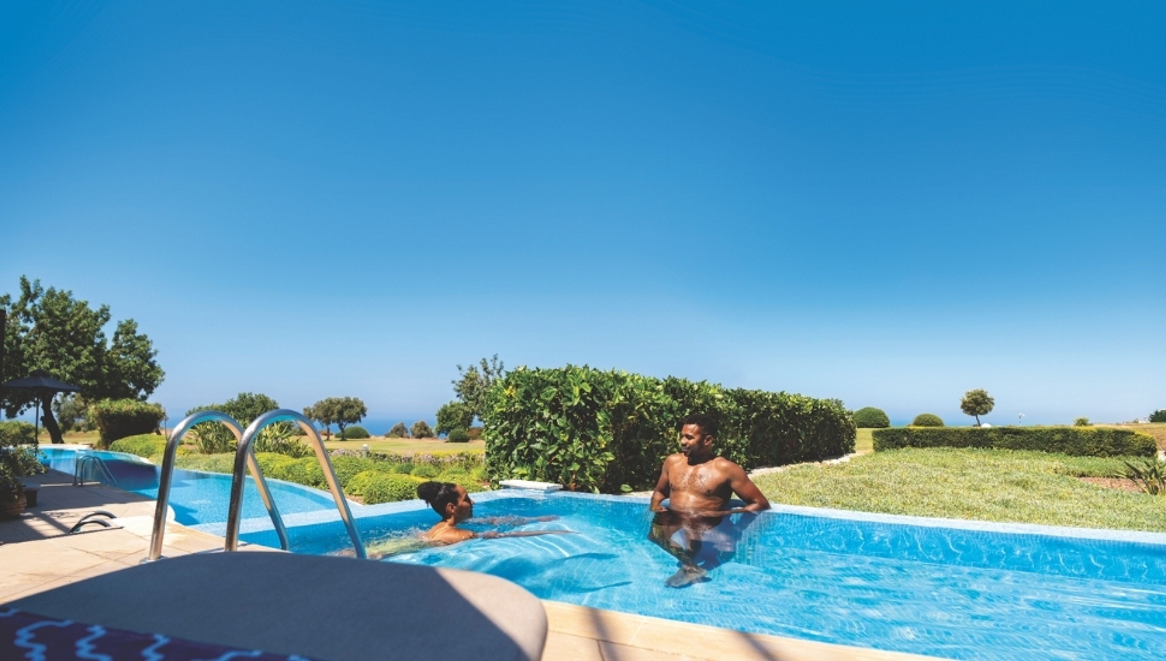 A man and a woman relax in an outdoor swimming pool under a clear blue sky, with green foliage and the sea in the background.