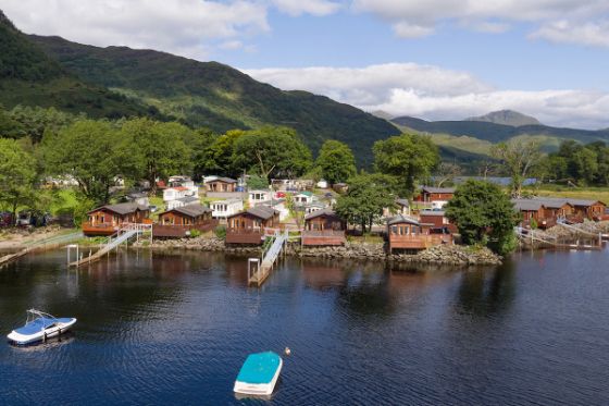 Waterfront wooden lodges and mobile homes with boats on a lake, backed by mountains.
