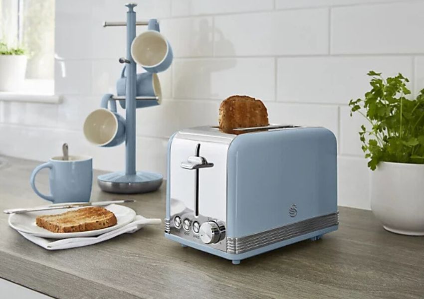 A light blue retro toaster with toast popping up on a kitchen counter, next to a mug tree holding matching cups and a plate with toast.