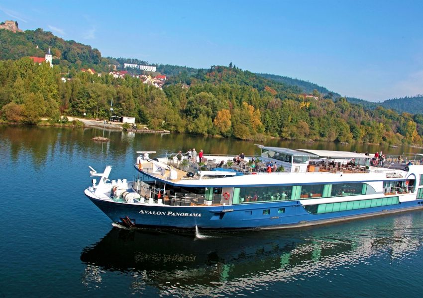 The Avalon Panorama river cruise ship sails on a river with a town and autumnal forest in the background.