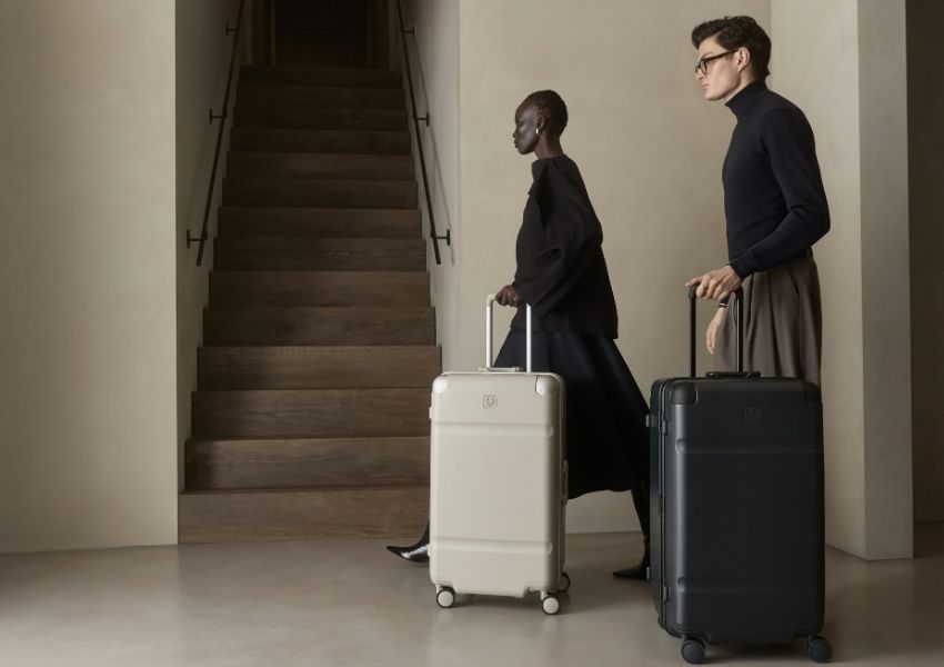 Two people pull a light and dark rolling suitcase past a staircase.