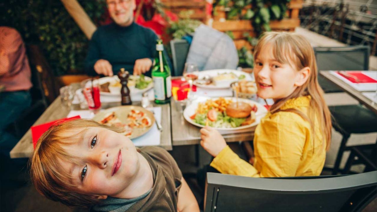 A family dines outdoors, with a smiling boy in the foreground and a girl smiling over her plate of food.