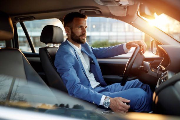 A man in a blue suit sits in the driver's seat of a car, lit by warm sunlight.