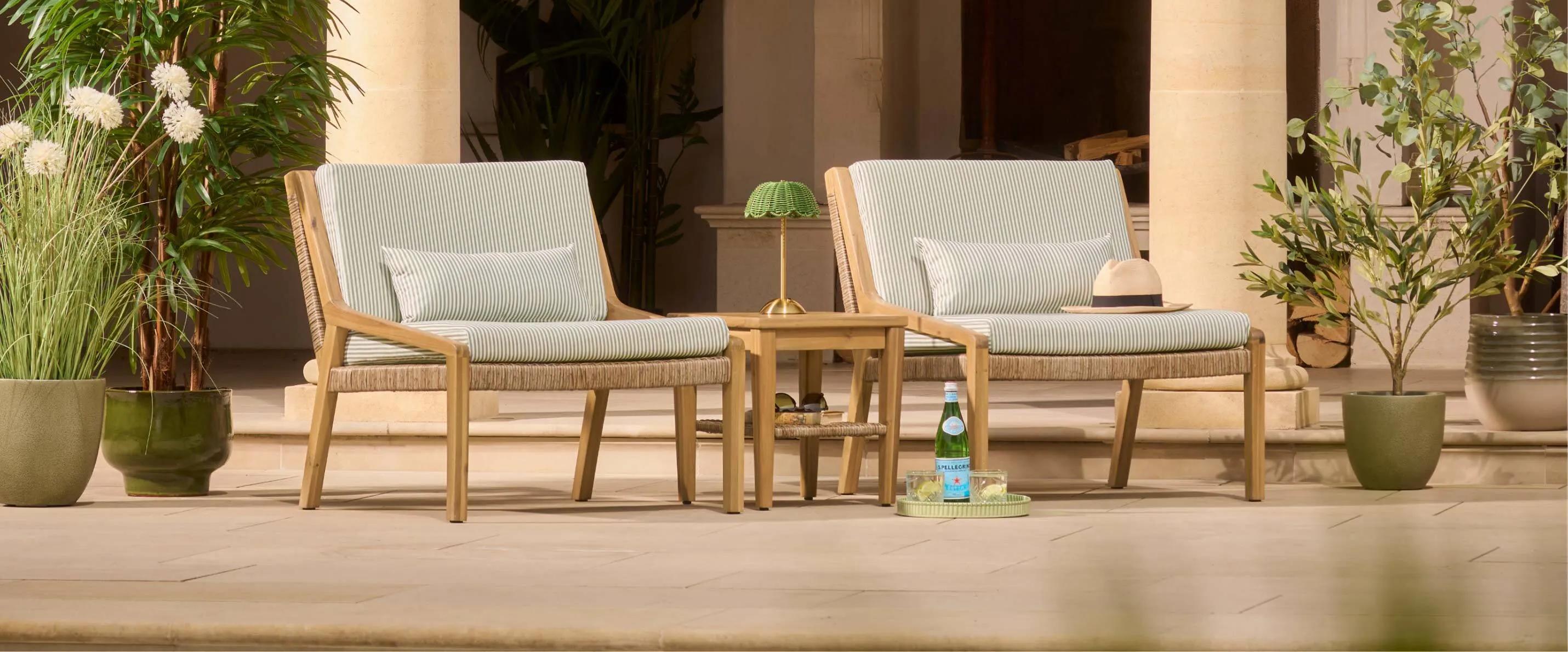 Two outdoor lounge chairs with light green striped cushions and a small wooden side table on a patio, surrounded by potted plants.