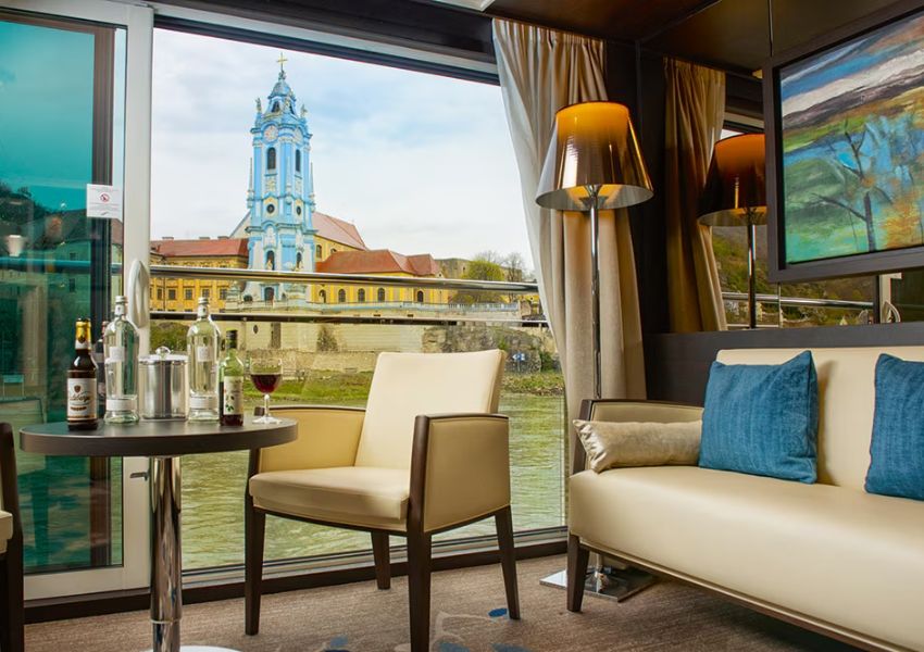 A lounge area with a panoramic window overlooking a river and the blue tower of Dürnstein Abbey, with a table of drinks, a chair, and a sofa.