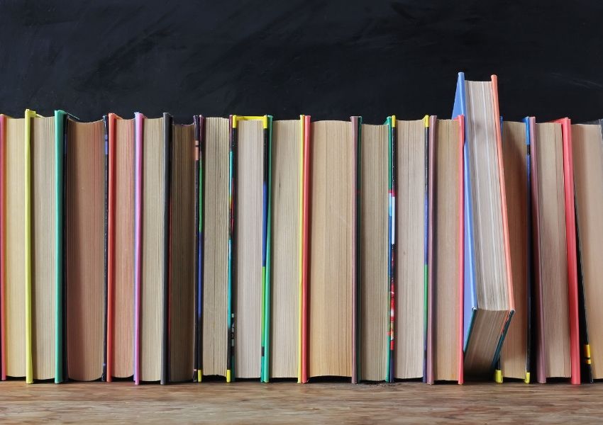 A row of colorful books on a wooden surface in front of a chalkboard, with one blue book angled out.