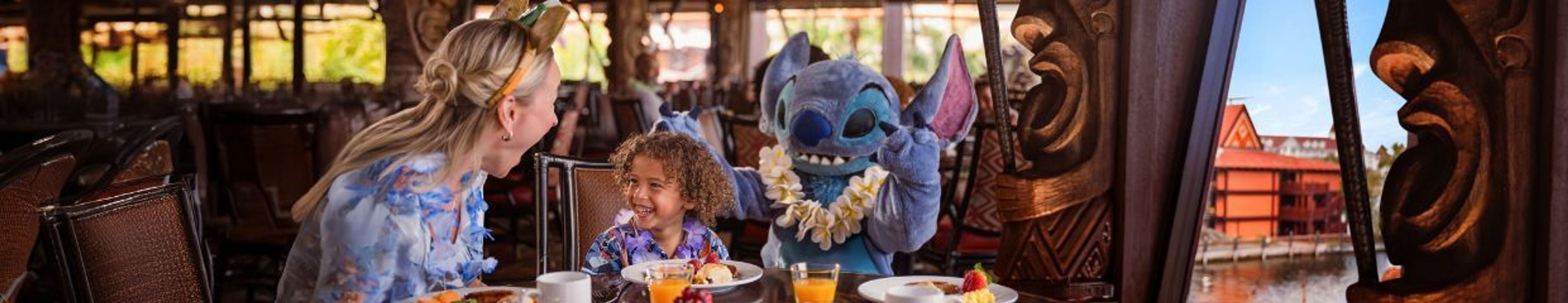 Stitch, wearing a lei, smiles with a woman and child at a breakfast table in a Polynesian-themed restaurant overlooking water.