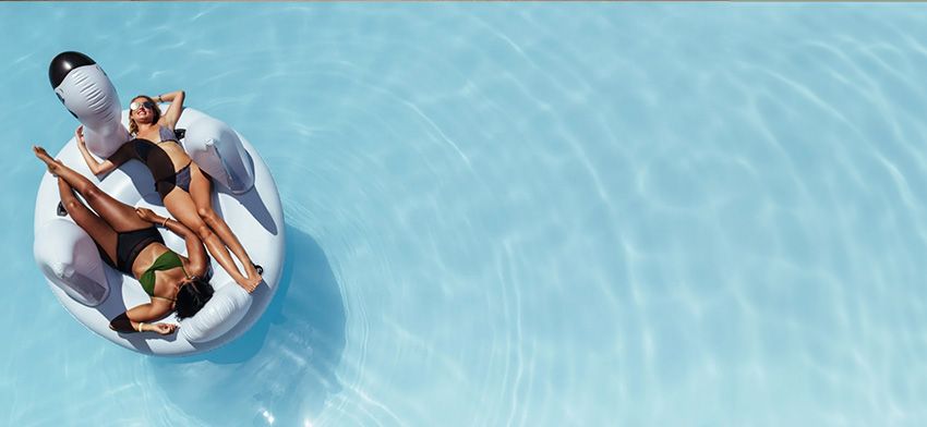 Two women sunbathing on a white swan float in a swimming pool.