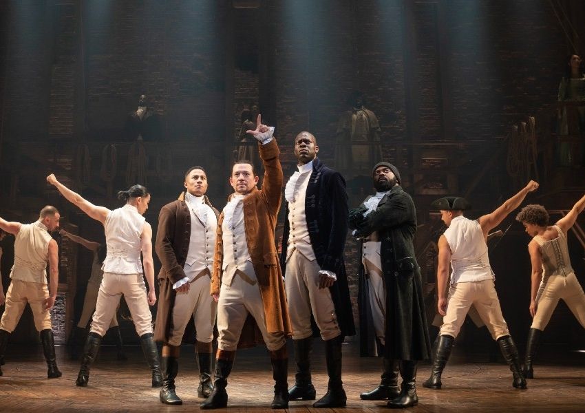 The cast of the musical Hamilton on stage, with a central actor in a brown coat raising his arm.