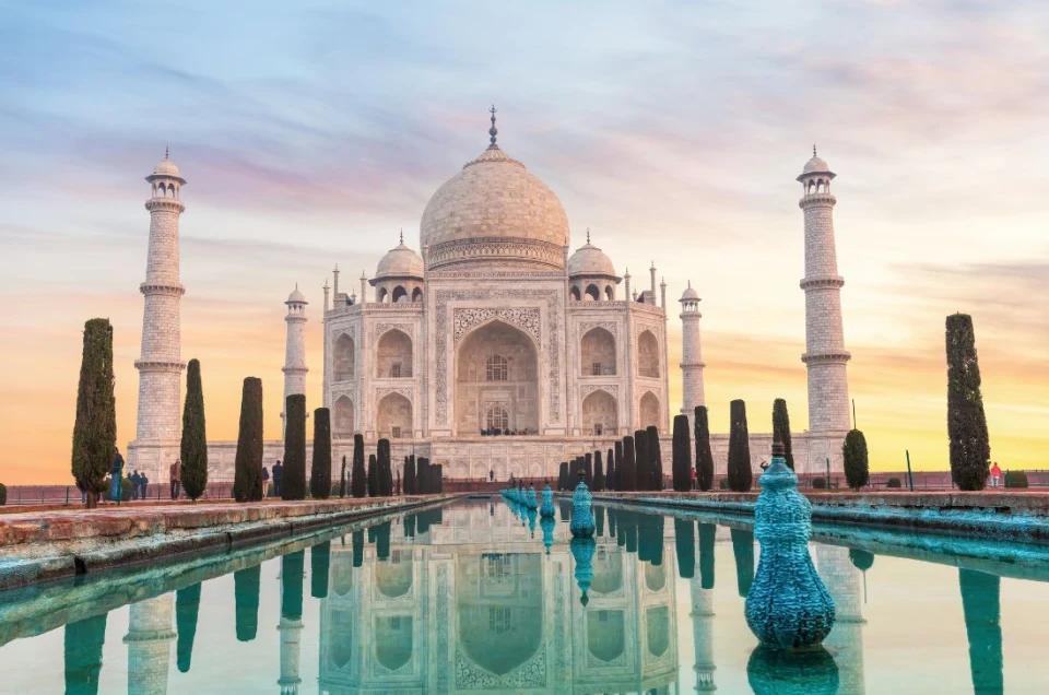 The white marble Taj Mahal reflected in a long pool of water under a colorful sunset sky.