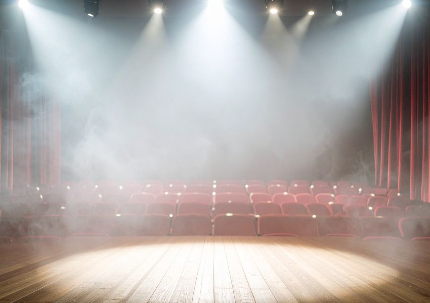 Empty theater stage with spotlights, fog, red curtains, and rows of red seats.