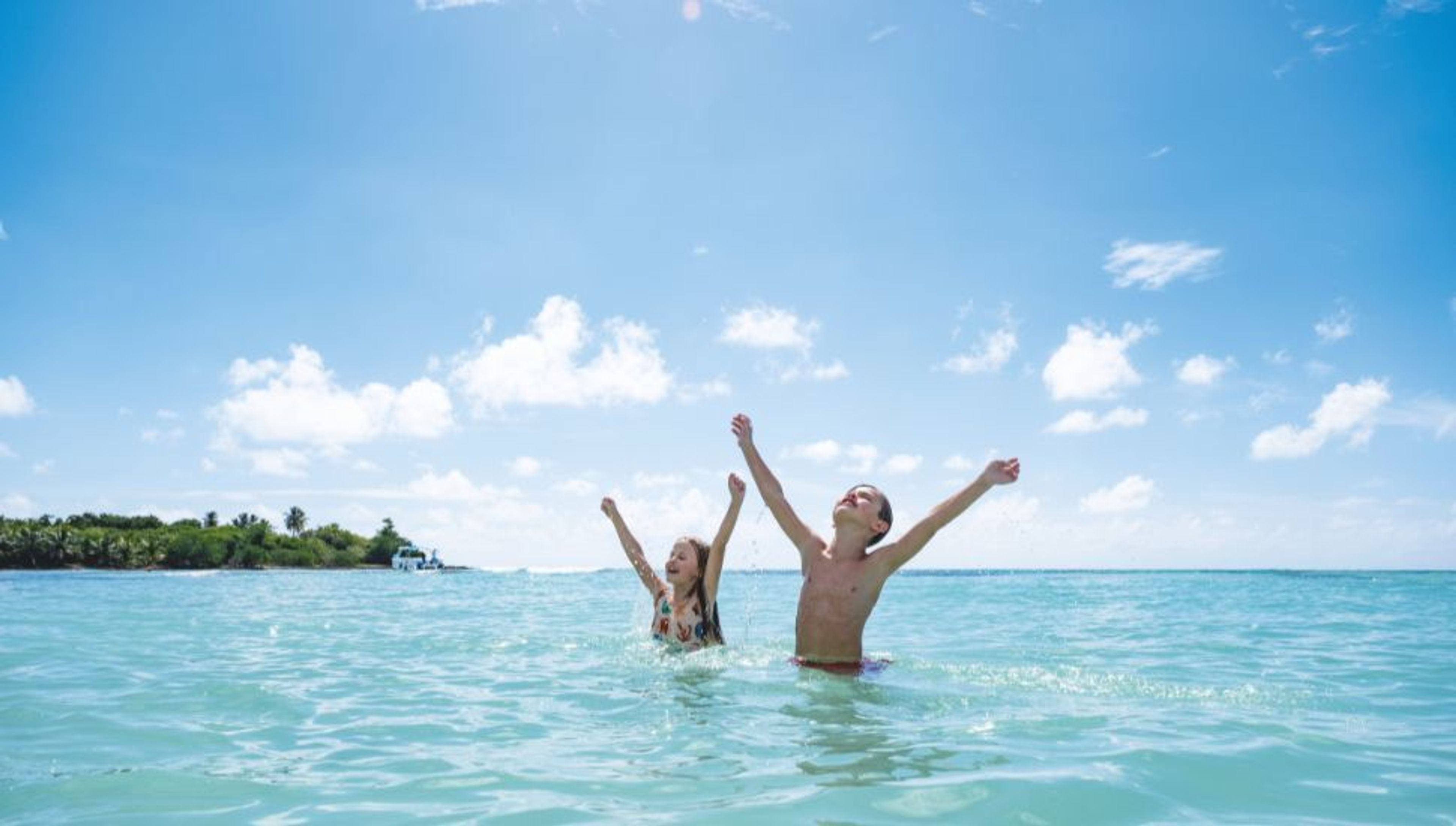 Two children raise their arms in clear blue ocean water on a sunny day.