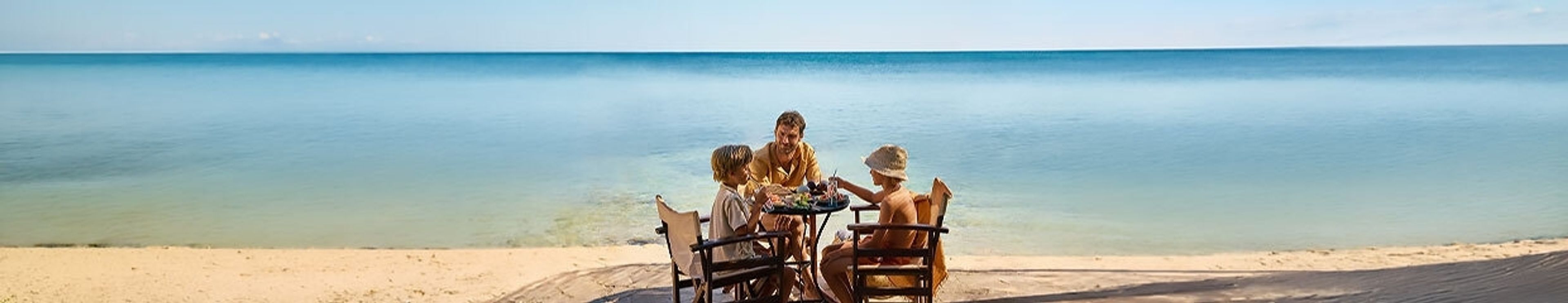 Three people sitting at a table on a sandy beach facing the calm blue ocean.