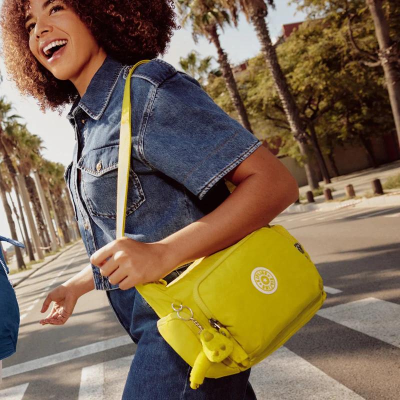 Smiling person with curly hair walks on a crosswalk, carrying a bright yellow crossbody bag with a monkey keychain.