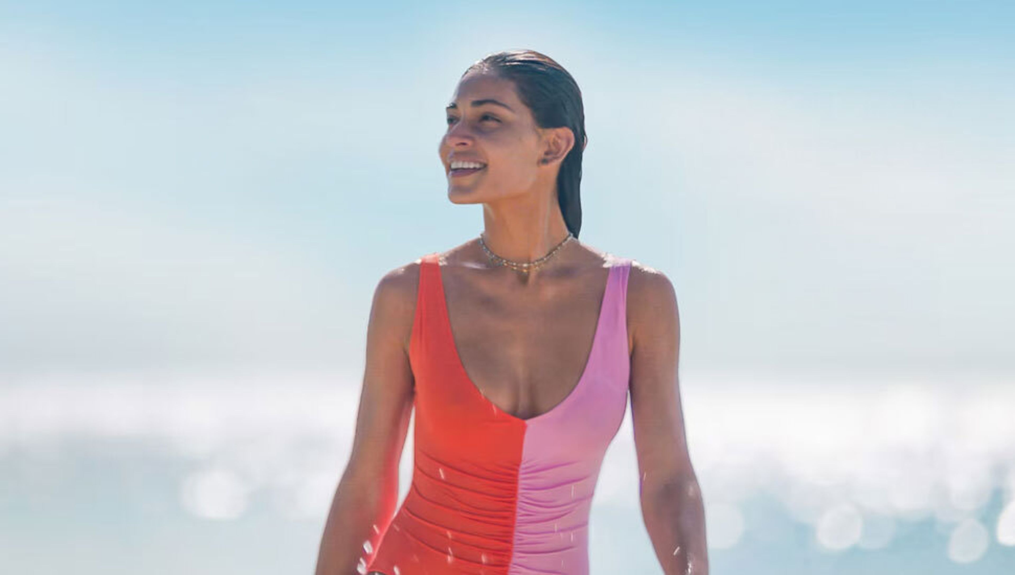 A smiling woman with wet hair in a red and pink swimsuit, looking left against a bright ocean background.