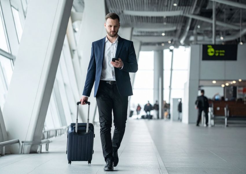 A businessman walks through an airport terminal, pulling a suitcase and looking at his phone.