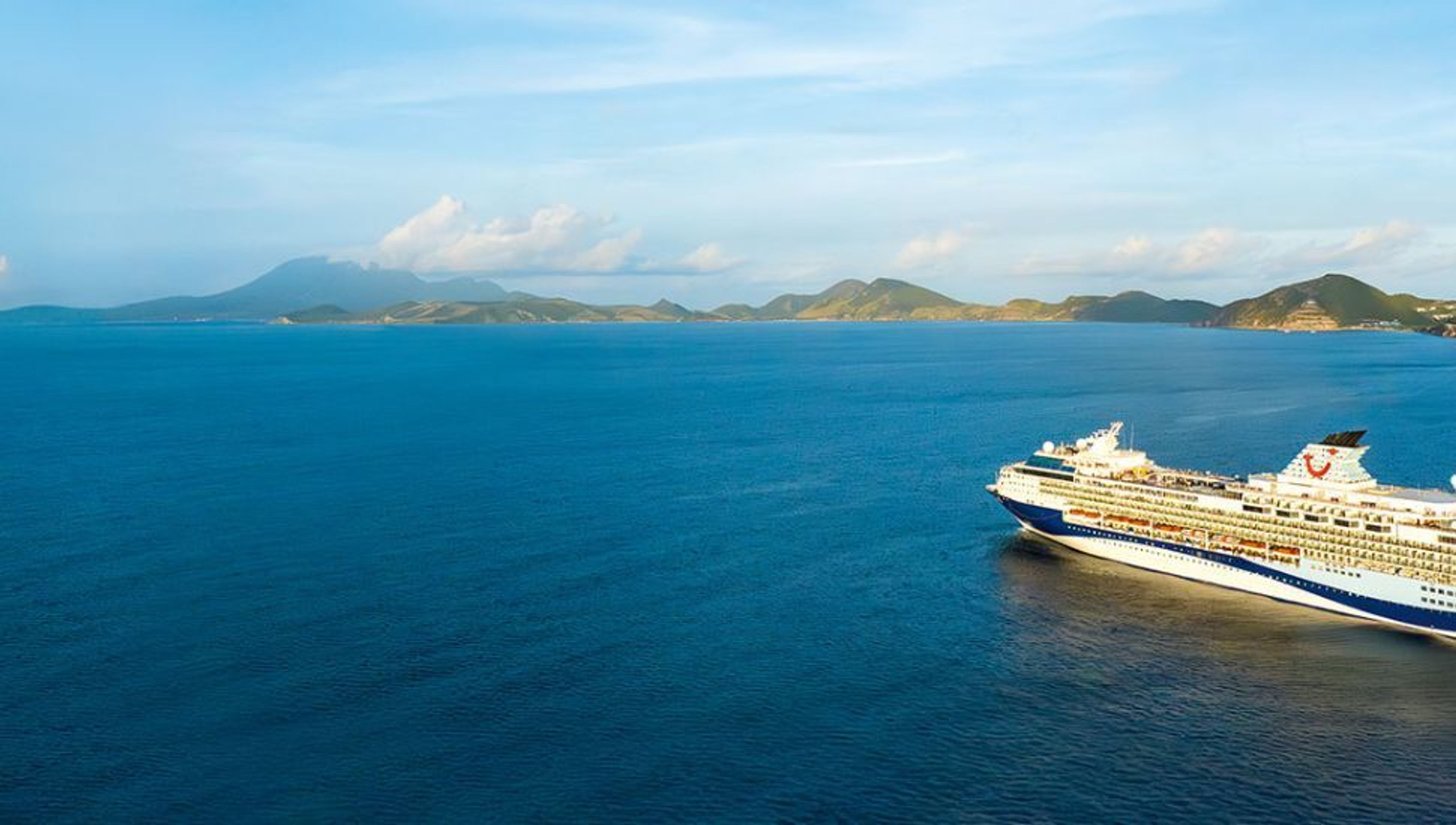 A cruise ship sails on blue water with mountainous islands in the background under a partly cloudy sky.