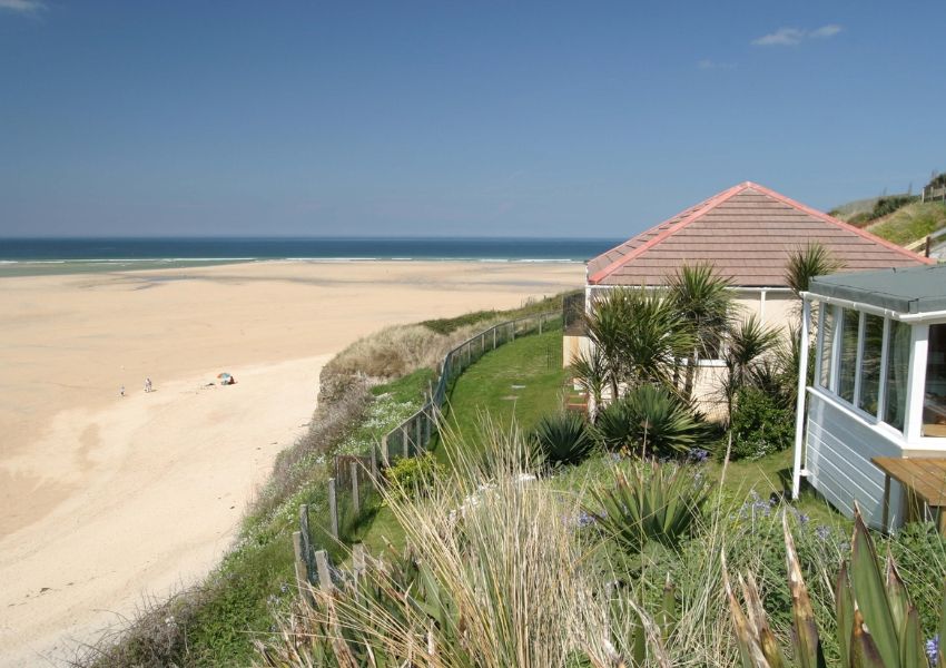 A house on a grassy cliff overlooking a wide sandy beach and ocean under a clear blue sky.