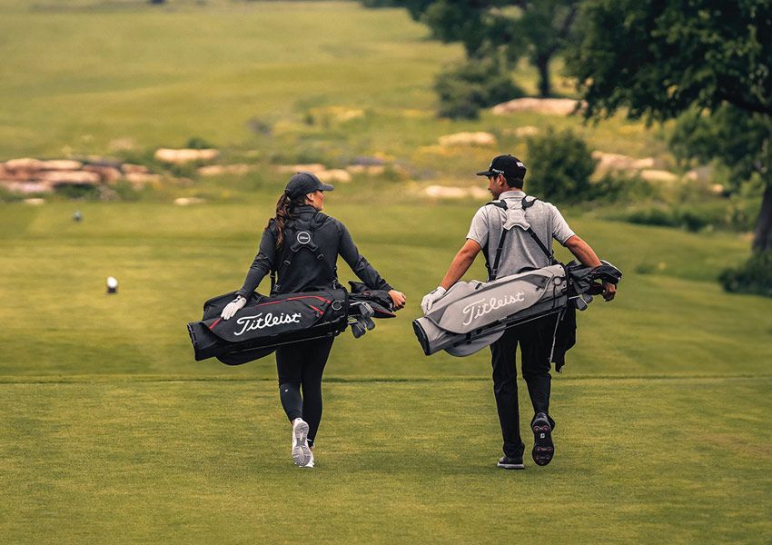 Two golfers, a woman and a man, walk across a green course carrying Titleist golf bags.