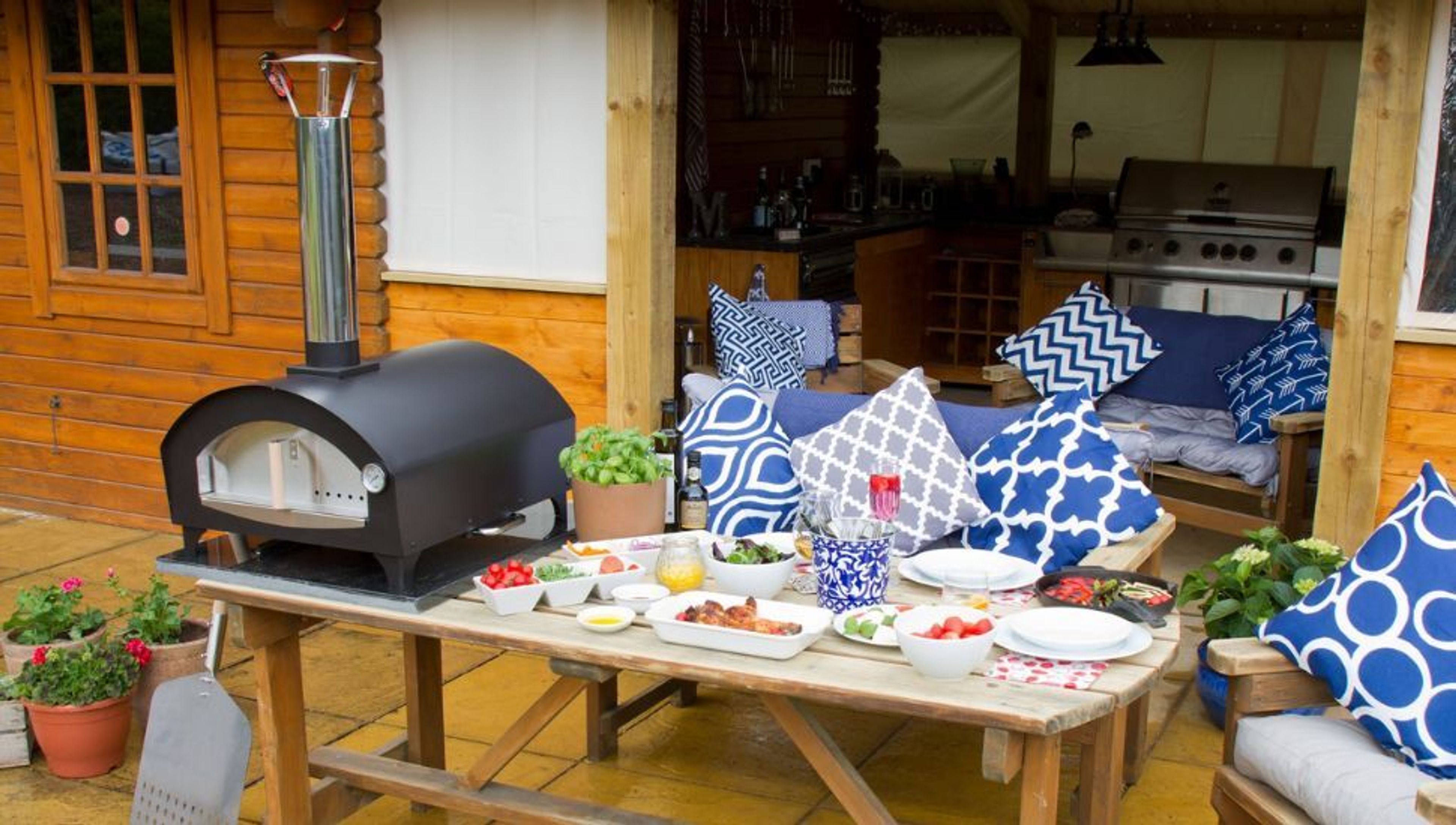 A vibrant outdoor living space featuring a black pizza oven on a wooden table set with food, and a seating area with blue and white cushions.