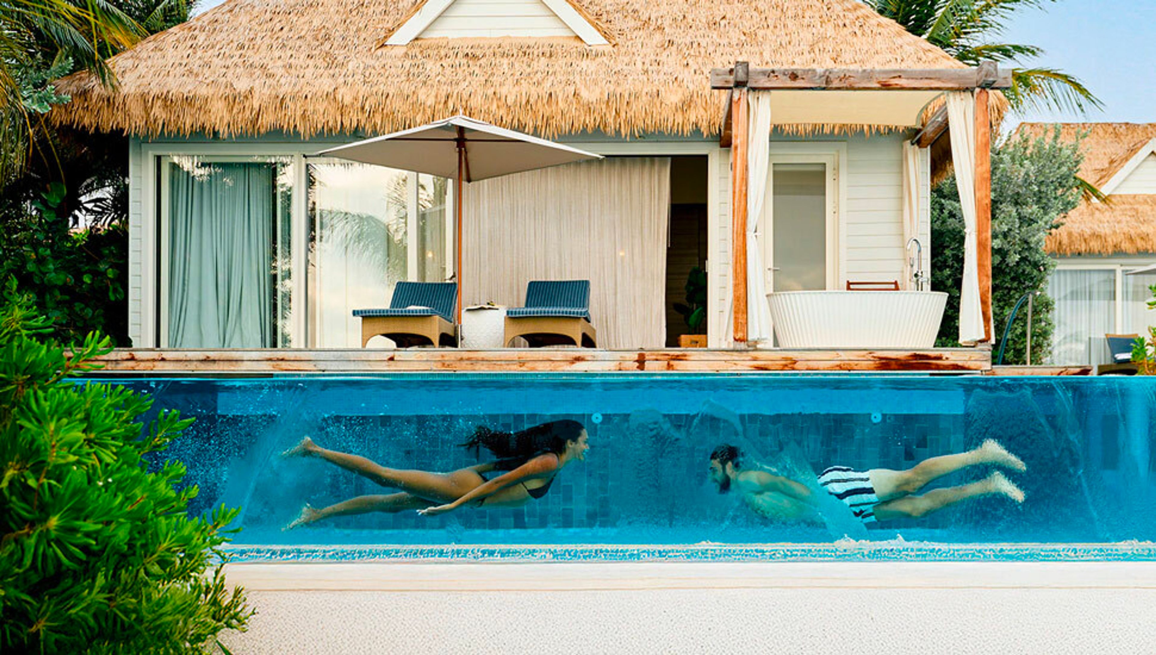 Two people swimming underwater in a glass-walled pool in front of a luxury thatched-roof villa.