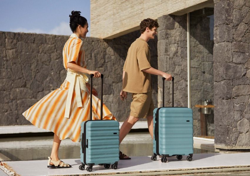 A man and woman walk with light blue suitcases beside a modern stone building.