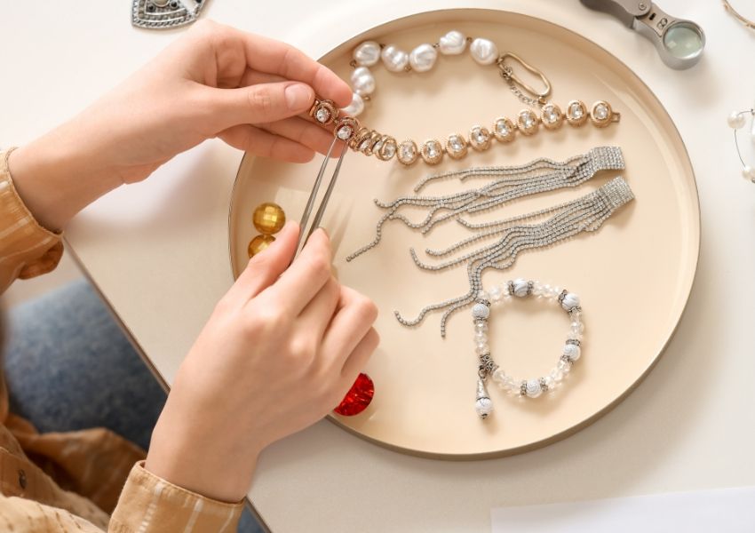 Hands using tweezers to work on a sparkling necklace on a tray with assorted jewelry.