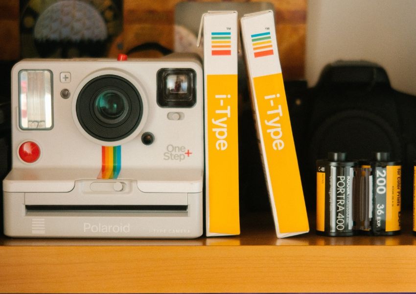 A white Polaroid OneStep+ camera with a rainbow stripe, two yellow i-Type film boxes, and several 35mm film rolls on a wooden shelf.