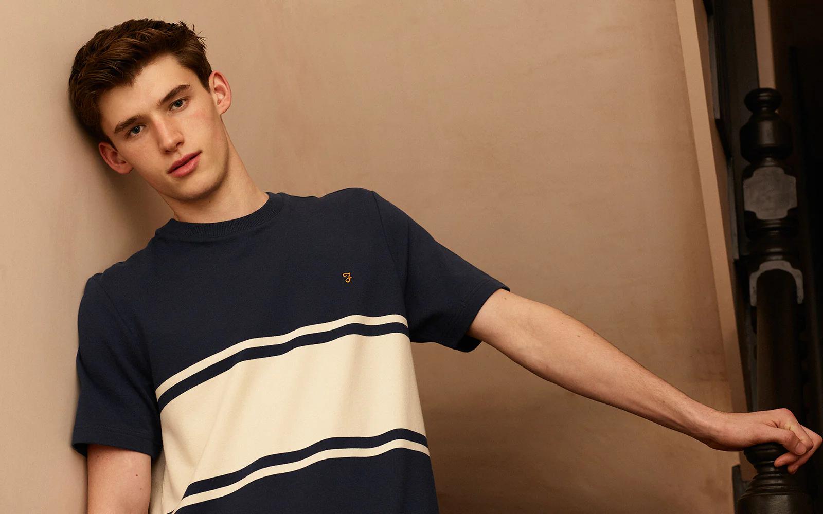 A young man in a navy blue and cream striped t-shirt leans against a wall, his hand resting on a dark banister.