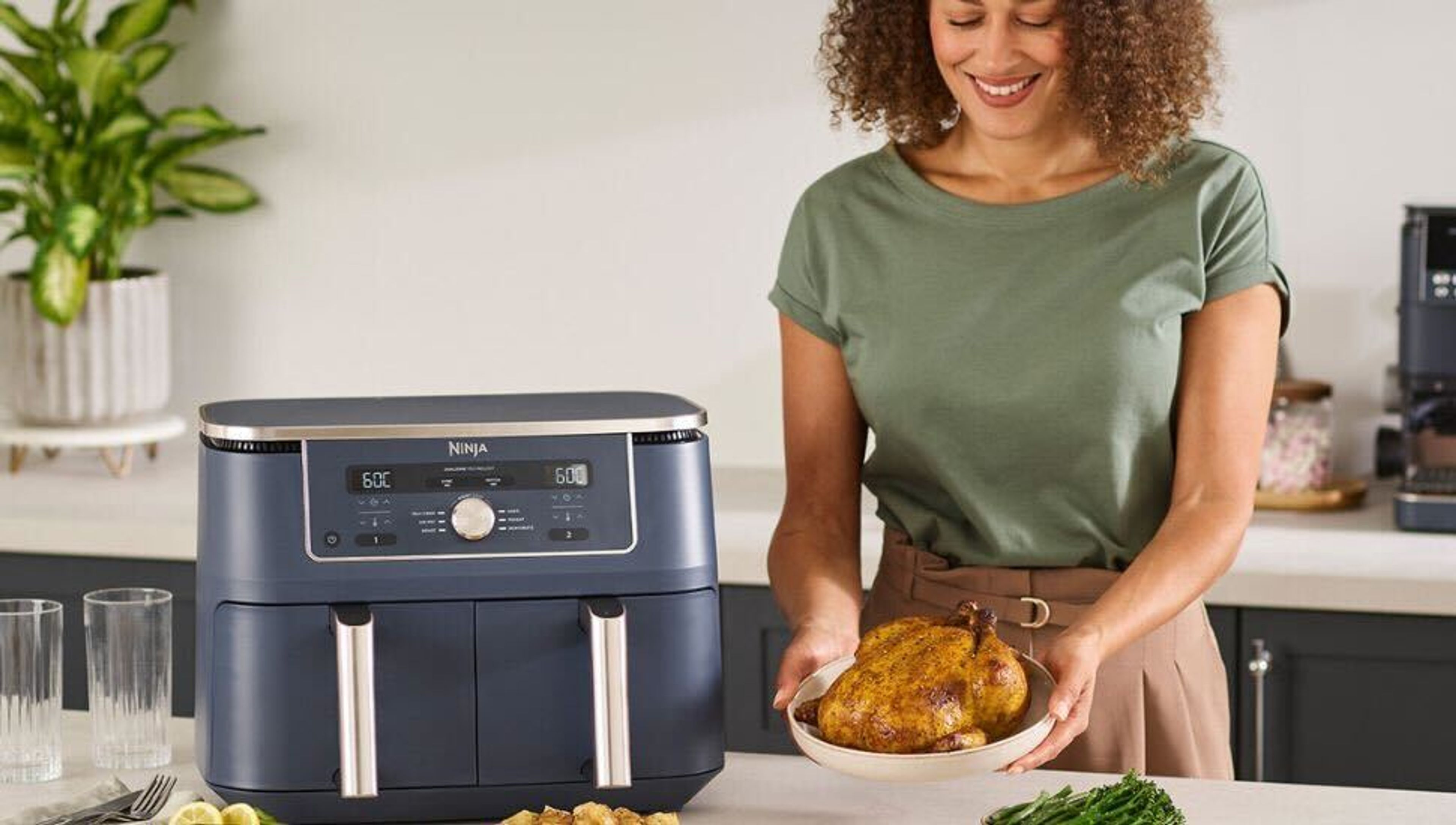 A woman holds a cooked chicken on a plate next to a dark blue Ninja DualZone air fryer in a kitchen.
