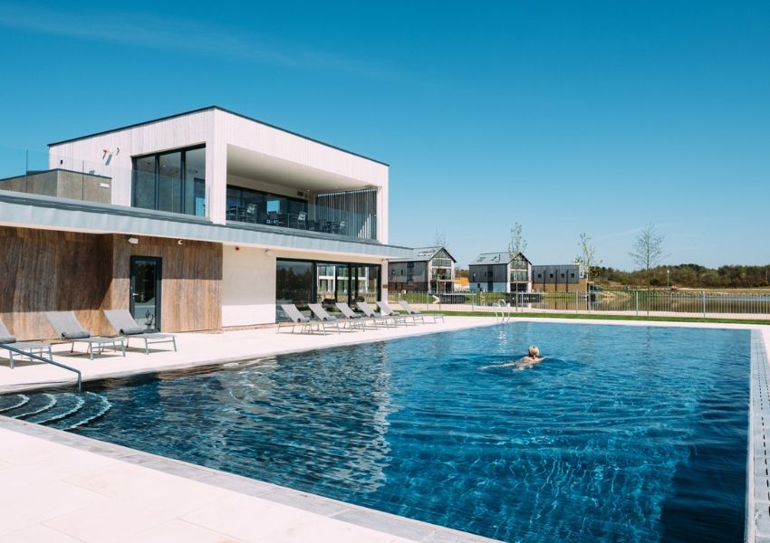 An outdoor swimming pool with a person swimming, a modern building, and houses by a lake under a blue sky.