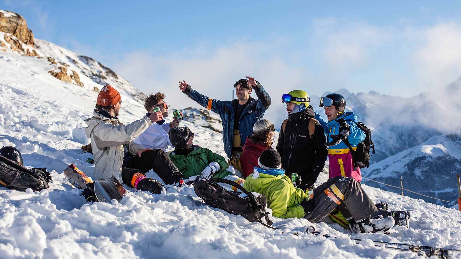 A group of skiers and snowboarders relaxing in the snow on a sunny mountain day, some toasting with drinks.