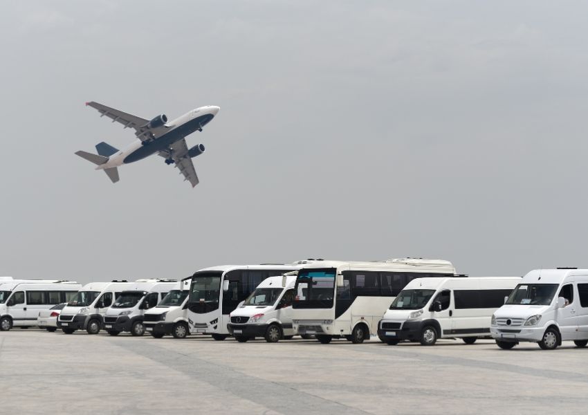 A white and blue airplane takes off over a row of white passenger vans and buses on a tarmac.