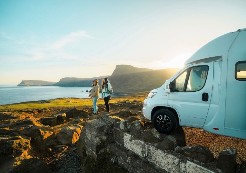 Two women and a camper van overlook a scenic coastal landscape at sunset.