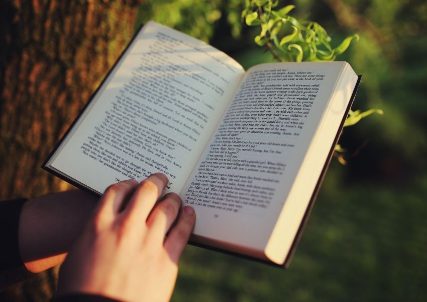 Hands holding an open book outdoors, illuminated by soft sunlight.