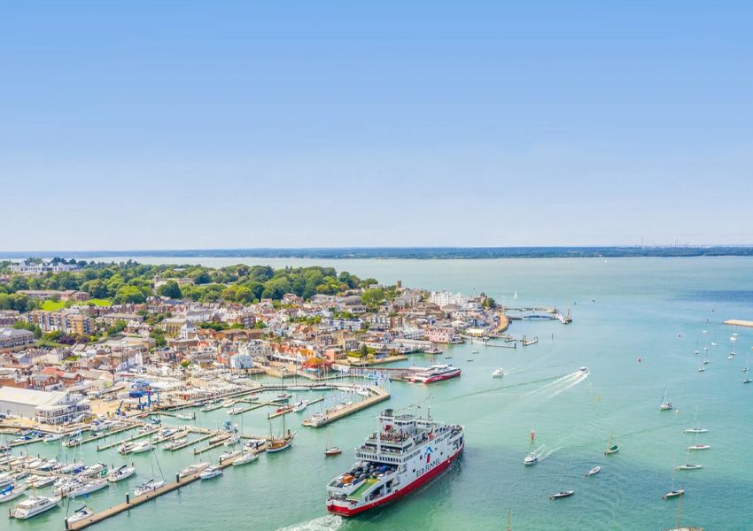 Aerial view of a coastal town and marina filled with boats, a large ferry, and clear blue water.