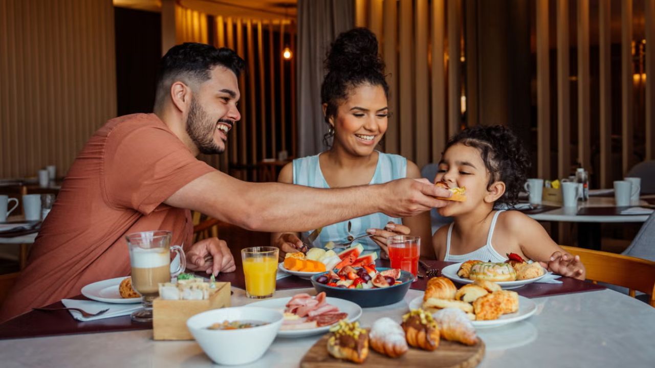 A man feeds his smiling daughter a bite of food during a family breakfast with a woman.