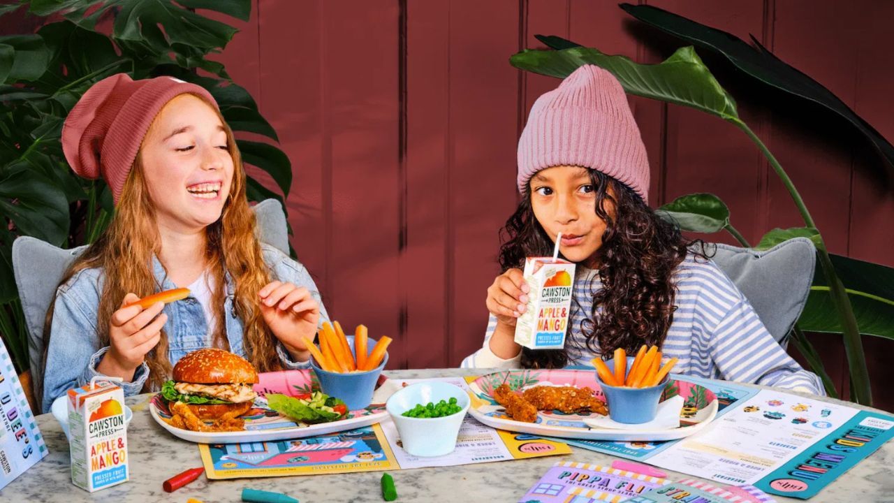 Two girls, one laughing with a burger and fries, the other sipping juice with chicken tenders and peas.