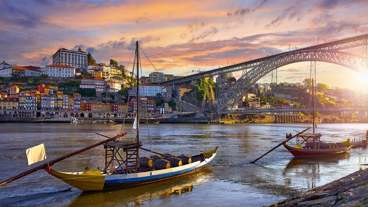 Sunset over Porto with traditional boats on the Douro River, colorful riverside buildings, and the Dom Luís I Bridge.